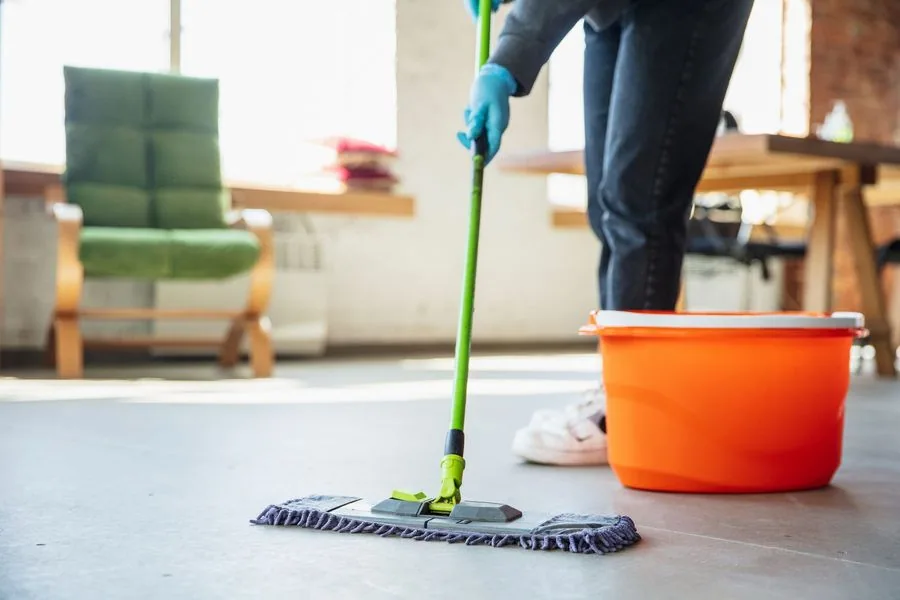 Person mopping a floor with a green mop and an orange bucket in a room with a green chair and a wooden table.
