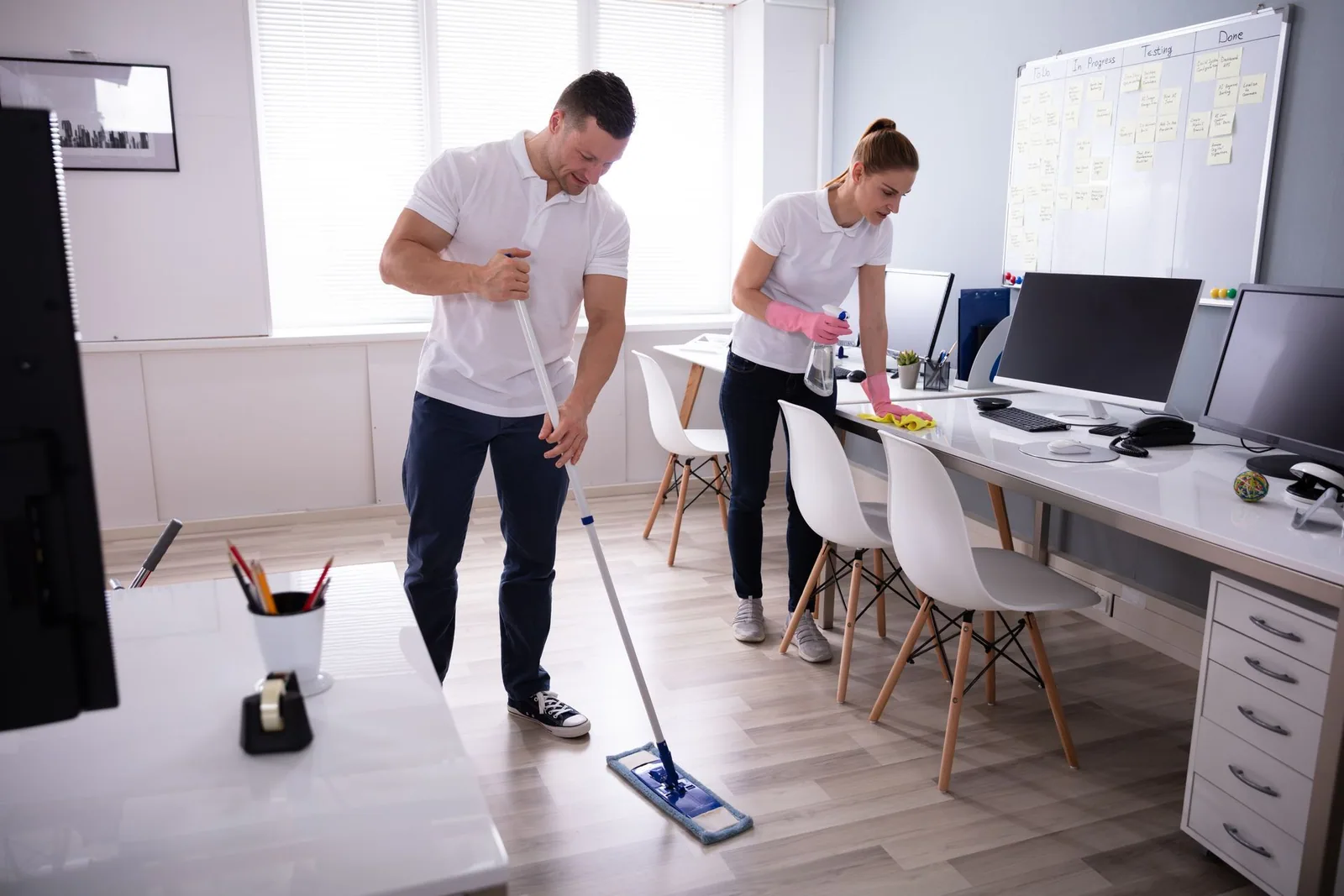Two people cleaning an office: man mopping floor, woman wiping desk.