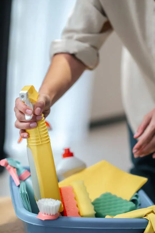 Person holding a yellow spray bottle, reaching into a blue cleaning supplies bucket.